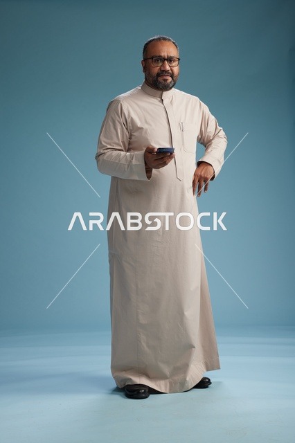 بورتريه لرجل  عربي سعودي خليجي يرتدي ثوب بيج ويحمل هاتفاً ذكياً, استخدام جهاز الجوال, كامل الجسم, خلفيه زرقاء