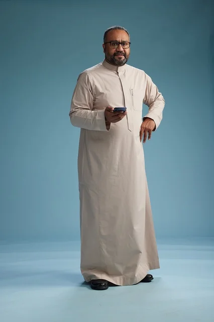 A portrait of an Arab Saudi Gulf man wearing a beige thobe and holding a smartphone, using a mobile device, full body, blue background.