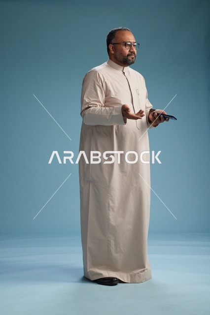 بورتريه لرجل  عربي سعودي خليجي يرتدي ثوب بيج ويحمل هاتفاً ذكياً, استخدام جهاز الجوال, كامل الجسم, خلفيه زرقاء