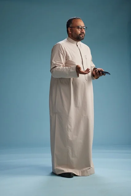 A portrait of an Arab Saudi Gulf man wearing a beige thobe and holding a smartphone, using a mobile device, full body, blue background.
