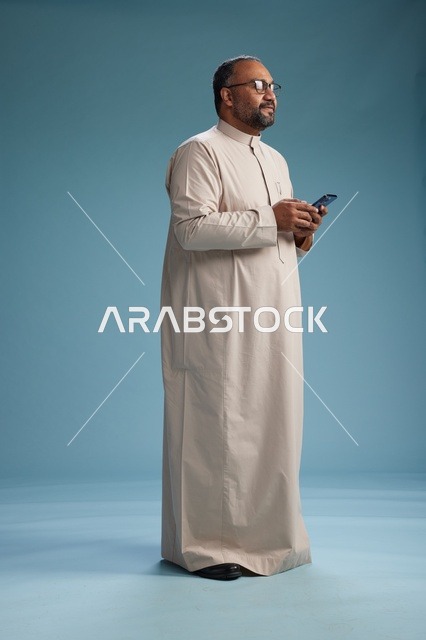 بورتريه لرجل  عربي سعودي خليجي يرتدي ثوب بيج ويحمل هاتفاً ذكياً, استخدام جهاز الجوال, كامل الجسم, خلفيه زرقاء