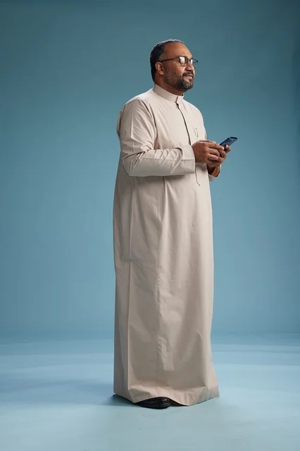 A portrait of an Arab Saudi Gulf man wearing a beige thobe and holding a smartphone, using a mobile device, full body, blue background.