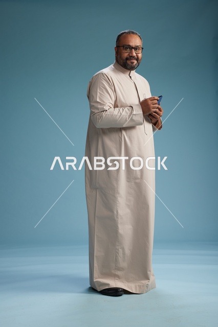 بورتريه لرجل  عربي سعودي خليجي يرتدي ثوب بيج ويحمل هاتفاً ذكياً, استخدام جهاز الجوال, كامل الجسم, خلفيه زرقاء