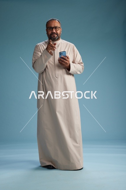 بورتريه لرجل  عربي سعودي خليجي يرتدي ثوب بيج ويحمل هاتفاً ذكياً, استخدام جهاز الجوال, كامل الجسم, خلفيه زرقاء