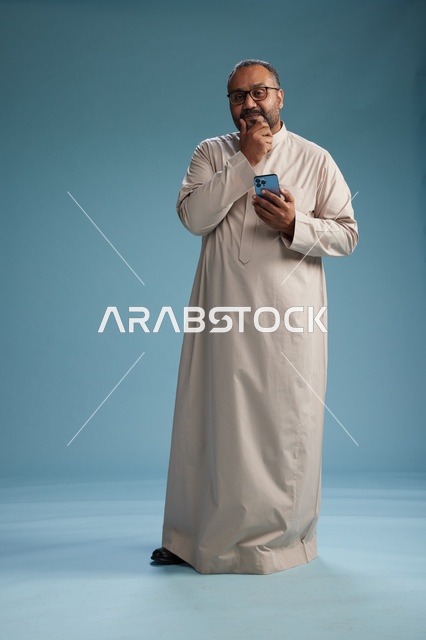 بورتريه لرجل  عربي سعودي خليجي يرتدي ثوب بيج ويحمل هاتفاً ذكياً, استخدام جهاز الجوال, كامل الجسم, خلفيه زرقاء