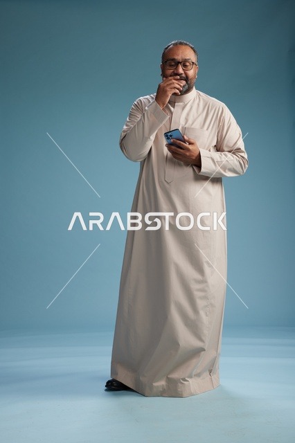 بورتريه لرجل  عربي سعودي خليجي يرتدي ثوب بيج ويحمل هاتفاً ذكياً, استخدام جهاز الجوال, كامل الجسم, خلفيه زرقاء