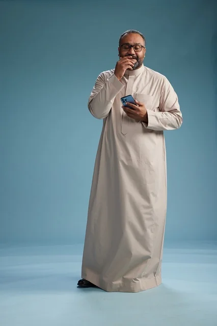 A portrait of an Arab Saudi Gulf man wearing a beige thobe and holding a smartphone, using a mobile device, full body, blue background.