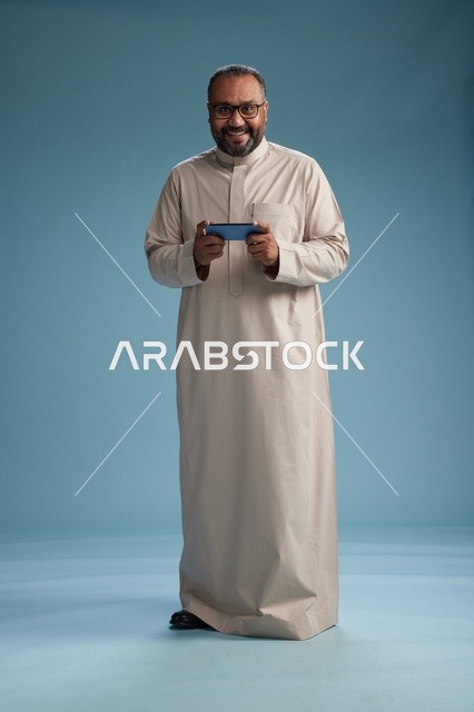 بورتريه لرجل  عربي سعودي خليجي يرتدي ثوب بيج ويحمل هاتفاً ذكياً, استخدام جهاز الجوال, كامل الجسم, خلفيه زرقاء