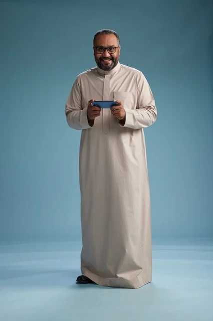 A portrait of an Arab Saudi Gulf man wearing a beige thobe and holding a smartphone, using a mobile device, full body, blue background.