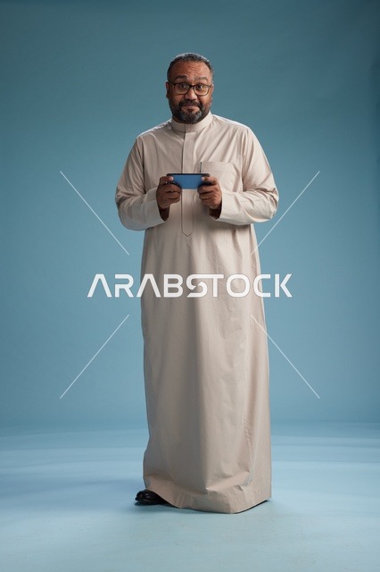 بورتريه لرجل  عربي سعودي خليجي يرتدي ثوب بيج ويحمل هاتفاً ذكياً, استخدام جهاز الجوال, كامل الجسم, خلفيه زرقاء