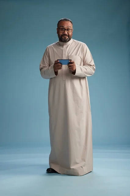 A portrait of an Arab Saudi Gulf man wearing a beige thobe and holding a smartphone, using a mobile device, full body, blue background.