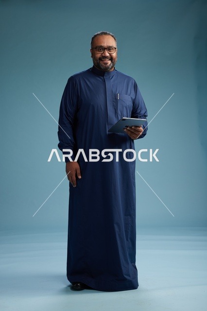 بورتريه لرجل عربي سعودي  خليجي بثوب أزرق يستخدم جهاز لوحي, استخدام التابليت المحمول, العمل اونلاين, الجسم بالكامل, خلفيه زرقاء