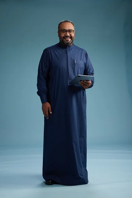 Portrait of a Saudi Arabian Gulf man in a blue thobe using a tablet, using a mobile tablet, working online, full body, blue background.