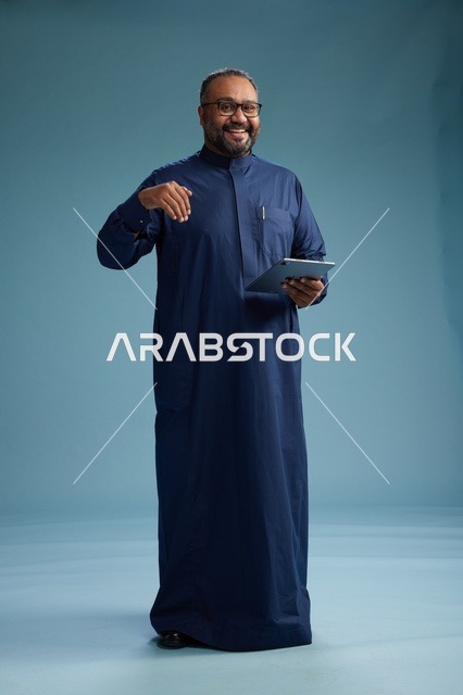 بورتريه لرجل عربي سعودي  خليجي بثوب أزرق يستخدم جهاز لوحي, استخدام التابليت المحمول, العمل اونلاين, الجسم بالكامل, خلفيه زرقاء