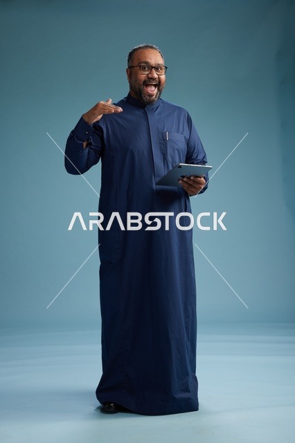 بورتريه لرجل عربي سعودي  خليجي بثوب أزرق يستخدم جهاز لوحي, استخدام التابليت المحمول, العمل اونلاين, الجسم بالكامل, خلفيه زرقاء