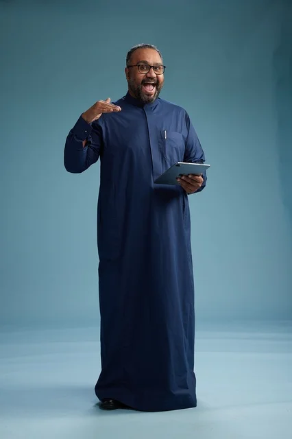 Portrait of a Saudi Arabian Gulf man in a blue thobe using a tablet, using a mobile tablet, working online, full body, blue background.