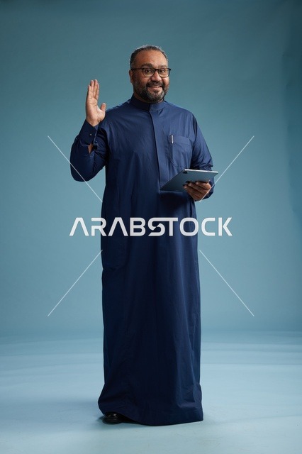 بورتريه لرجل عربي سعودي  خليجي بثوب أزرق يستخدم جهاز لوحي, استخدام التابليت المحمول, العمل اونلاين, الجسم بالكامل, خلفيه زرقاء
