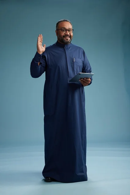 Portrait of a Saudi Arabian Gulf man in a blue thobe using a tablet, using a mobile tablet, working online, full body, blue background.