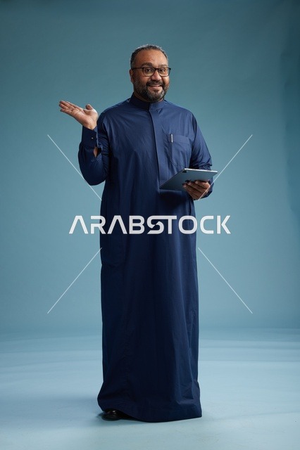 بورتريه لرجل عربي سعودي  خليجي بثوب أزرق يستخدم جهاز لوحي, استخدام التابليت المحمول, العمل اونلاين, الجسم بالكامل, خلفيه زرقاء