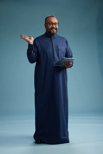 بورتريه لرجل عربي سعودي  خليجي بثوب أزرق يستخدم جهاز لوحي, استخدام التابليت المحمول, العمل اونلاين, الجسم بالكامل, خلفيه زرقاء
