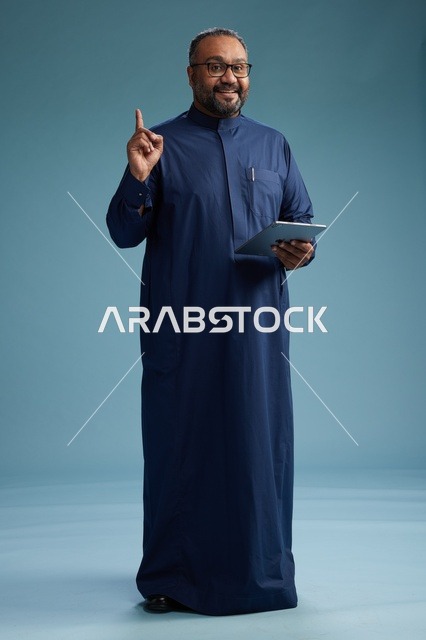 بورتريه لرجل عربي سعودي  خليجي بثوب أزرق يستخدم جهاز لوحي, استخدام التابليت المحمول, العمل اونلاين, الجسم بالكامل, خلفيه زرقاء