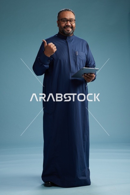 بورتريه لرجل عربي سعودي  خليجي بثوب أزرق يستخدم جهاز لوحي, استخدام التابليت المحمول, العمل اونلاين, الجسم بالكامل, خلفيه زرقاء