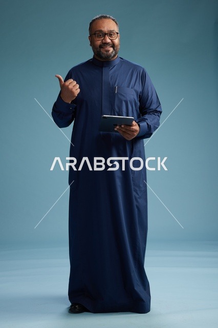 بورتريه لرجل عربي سعودي  خليجي بثوب أزرق يستخدم جهاز لوحي, استخدام التابليت المحمول, العمل اونلاين, الجسم بالكامل, خلفيه زرقاء