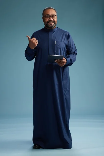 Portrait of a Saudi Arabian Gulf man in a blue thobe using a tablet, using a mobile tablet, working online, full body, blue background.