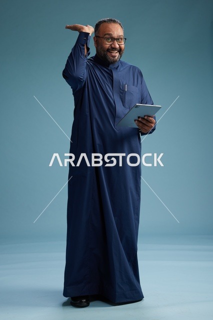 بورتريه لرجل عربي سعودي  خليجي بثوب أزرق يستخدم جهاز لوحي, استخدام التابليت المحمول, العمل اونلاين, الجسم بالكامل, خلفيه زرقاء