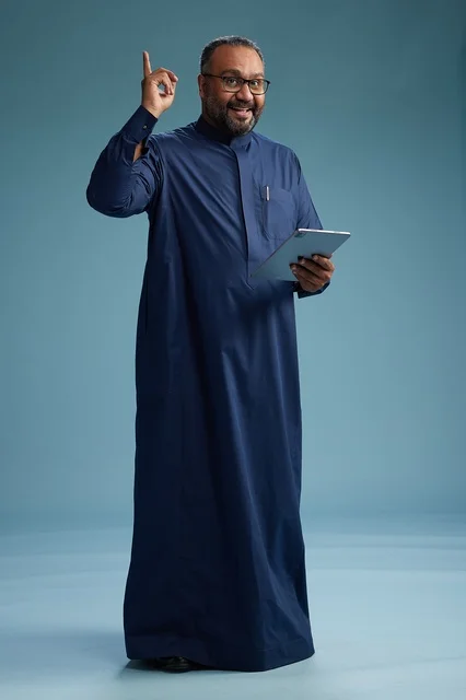 Portrait of a Saudi Arabian Gulf man in a blue thobe using a tablet, using a mobile tablet, working online, full body, blue background.