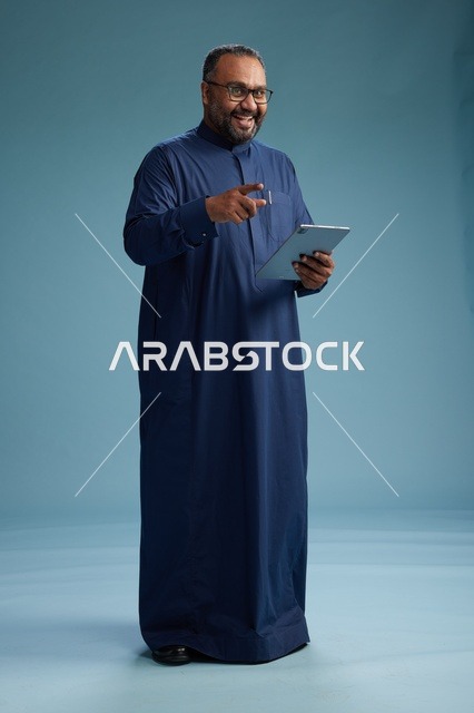Portrait of a Saudi Arabian Gulf man in a blue thobe using a tablet, using a mobile tablet, working online, full body, blue background.