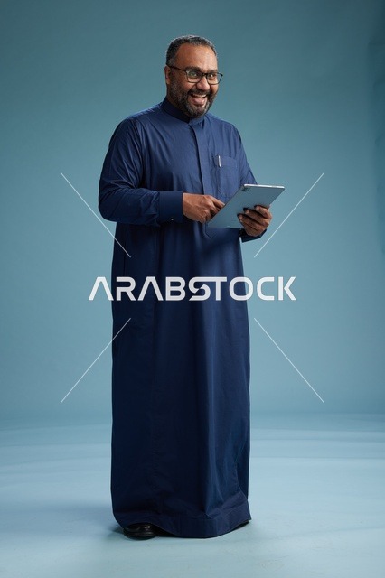 بورتريه لرجل عربي سعودي  خليجي بثوب أزرق يستخدم جهاز لوحي, استخدام التابليت المحمول, العمل اونلاين, الجسم بالكامل, خلفيه زرقاء