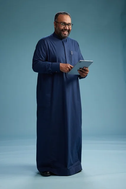 Portrait of a Saudi Arabian Gulf man in a blue thobe using a tablet, using a mobile tablet, working online, full body, blue background.