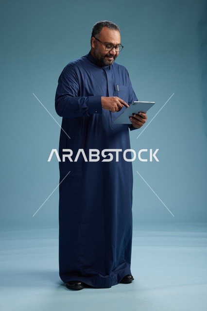 بورتريه لرجل عربي سعودي  خليجي بثوب أزرق يستخدم جهاز لوحي, استخدام التابليت المحمول, العمل اونلاين, الجسم بالكامل, خلفيه زرقاء