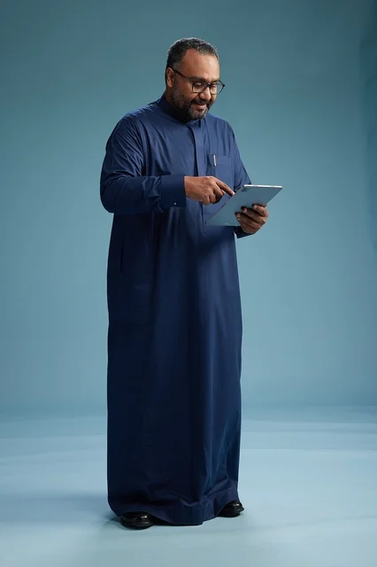 Portrait of a Saudi Arabian Gulf man in a blue thobe using a tablet, using a mobile tablet, working online, full body, blue background.