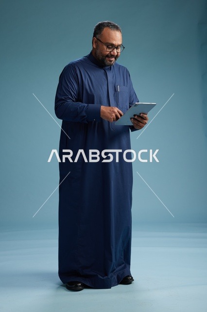 بورتريه لرجل عربي سعودي  خليجي بثوب أزرق يستخدم جهاز لوحي, استخدام التابليت المحمول, العمل اونلاين, الجسم بالكامل, خلفيه زرقاء