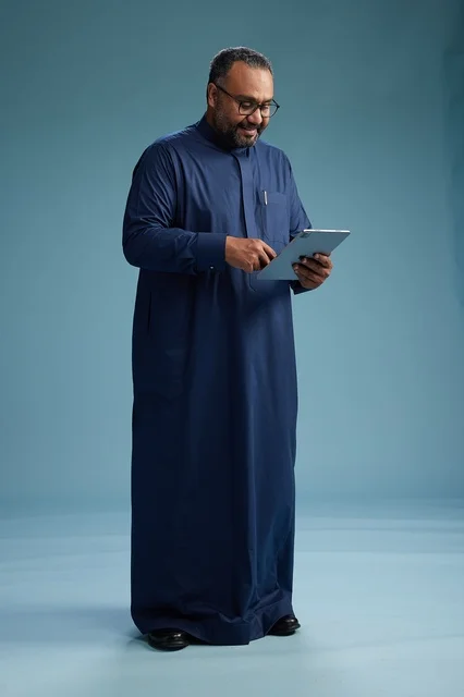 Portrait of a Saudi Arabian Gulf man in a blue thobe using a tablet, using a mobile tablet, working online, full body, blue background.