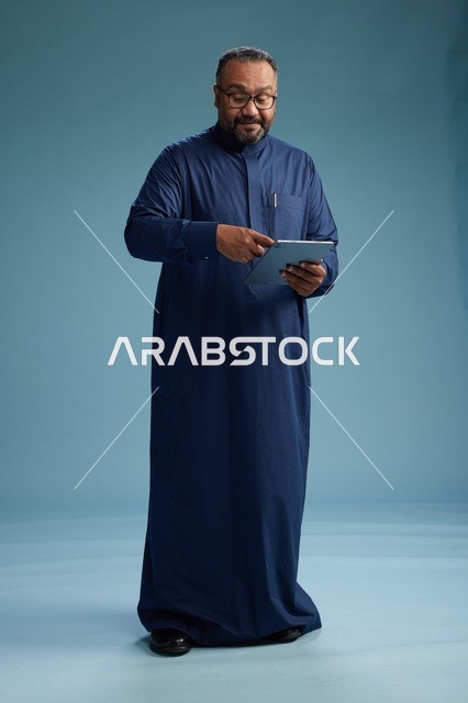بورتريه لرجل عربي سعودي  خليجي بثوب أزرق يستخدم جهاز لوحي, استخدام التابليت المحمول, العمل اونلاين, الجسم بالكامل, خلفيه زرقاء