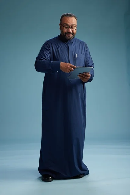 بورتريه لرجل عربي سعودي  خليجي بثوب أزرق يستخدم جهاز لوحي, استخدام التابليت المحمول, العمل اونلاين, الجسم بالكامل, خلفيه زرقاء