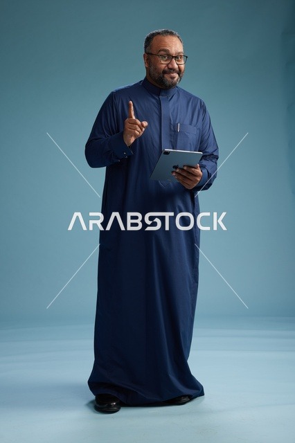 بورتريه لرجل عربي سعودي  خليجي بثوب أزرق يستخدم جهاز لوحي, استخدام التابليت المحمول, العمل اونلاين, الجسم بالكامل, خلفيه زرقاء