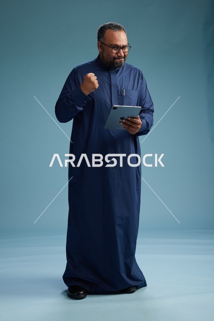بورتريه لرجل عربي سعودي  خليجي بثوب أزرق يستخدم جهاز لوحي, استخدام التابليت المحمول, العمل اونلاين, الجسم بالكامل, خلفيه زرقاء