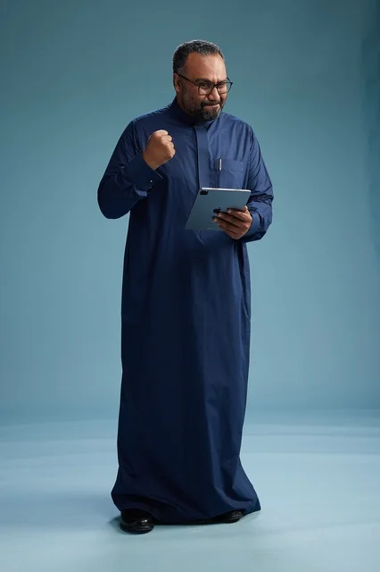 Portrait of a Saudi Arabian Gulf man in a blue thobe using a tablet, using a mobile tablet, working online, full body, blue background. Portrait of a Saudi Arabian Gulf man in a blue thobe using a tablet, using a mobile tablet, working online, full body, blue background.