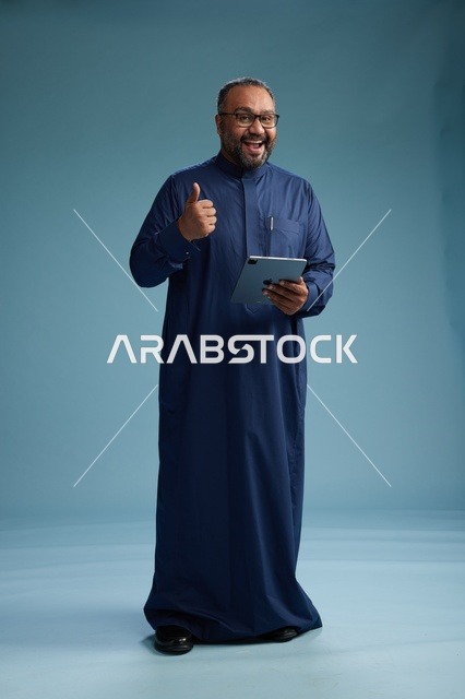 بورتريه لرجل عربي سعودي  خليجي بثوب أزرق يستخدم جهاز لوحي, استخدام التابليت المحمول, العمل اونلاين, الجسم بالكامل, خلفيه زرقاء