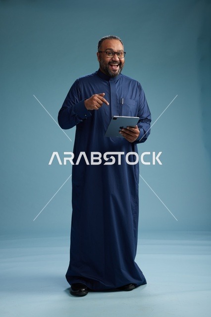 بورتريه لرجل عربي سعودي  خليجي بثوب أزرق يستخدم جهاز لوحي, استخدام التابليت المحمول, العمل اونلاين, الجسم بالكامل, خلفيه زرقاء