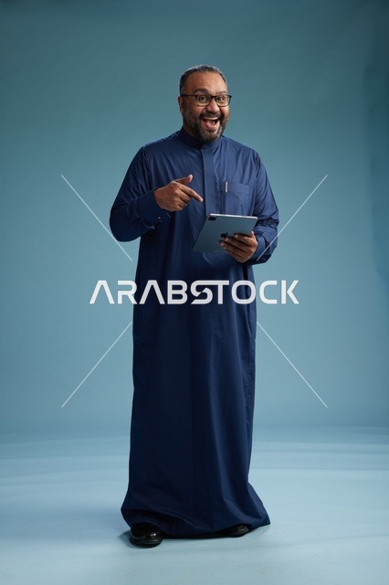 بورتريه لرجل عربي سعودي  خليجي بثوب أزرق يستخدم جهاز لوحي, استخدام التابليت المحمول, العمل اونلاين, الجسم بالكامل, خلفيه زرقاء