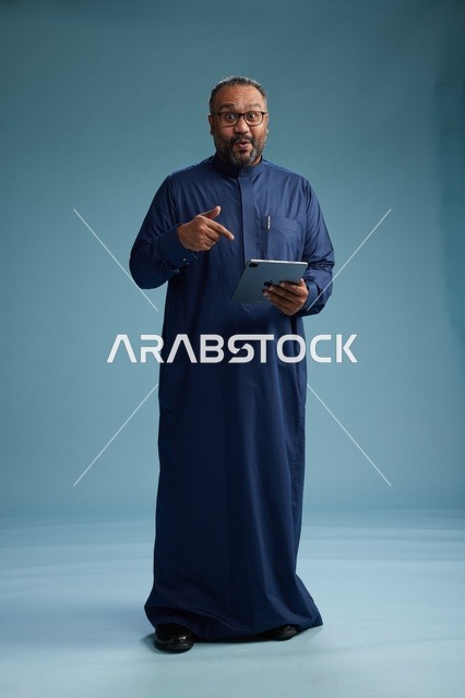 بورتريه لرجل عربي سعودي  خليجي بثوب أزرق يستخدم جهاز لوحي, استخدام التابليت المحمول, العمل اونلاين, الجسم بالكامل, خلفيه زرقاء