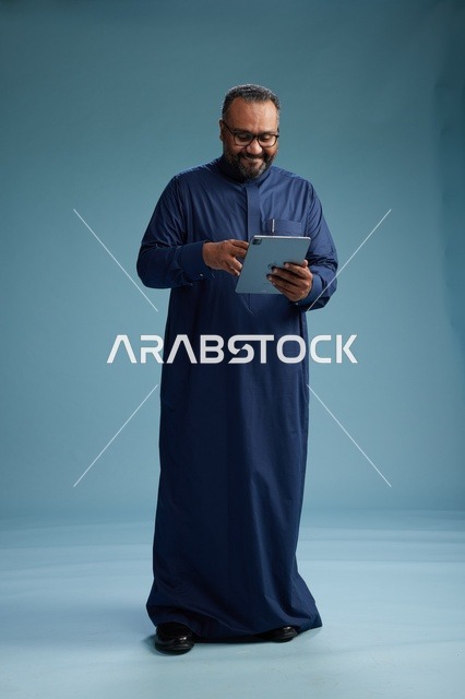بورتريه لرجل عربي سعودي  خليجي بثوب أزرق يستخدم جهاز لوحي, استخدام التابليت المحمول, العمل اونلاين, الجسم بالكامل, خلفيه زرقاء
