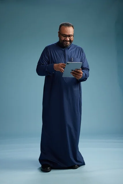 Portrait of a Saudi Arabian Gulf man in a blue thobe using a tablet, using a mobile tablet, working online, full body, blue background.