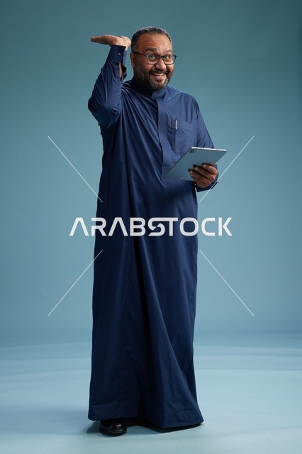 بورتريه لرجل عربي سعودي  خليجي بثوب أزرق يستخدم جهاز لوحي, استخدام التابليت المحمول, العمل اونلاين, الجسم بالكامل, خلفيه زرقاء