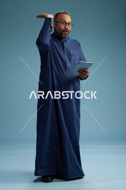 بورتريه لرجل عربي سعودي  خليجي بثوب أزرق يستخدم جهاز لوحي, استخدام التابليت المحمول, العمل اونلاين, الجسم بالكامل, خلفيه زرقاء